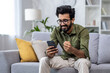 © Liubomir - Happy and satisfied with achievement results hispanic at home reading online win notification in online application on phone, man smiling holding hand up triumph gesture sitting sofa in living room.