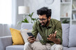 © Liubomir - Upset and sad man sitting on sofa at home in living room, hindu man reading bad news online from phone, depressed and sad man.