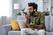 © Liubomir - Angry and nervous man talking on the phone sitting on the sofa in the living room, hispanic man yelling at the interlocutor on the smartphone, dissatisfied with the service, the customer complains.