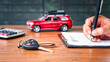© Boxyray - Car loan or buying a new car concept. Car key on table with a man signing on leasing or document  form and red car in background. Leasing and insurance business.