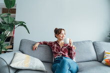 Young Smiling Woman Sitting On Sofa And Looking Away While Drinking Coffee Or Tea. Young Brunette Woman Relaxing After Housekeeping, Home Cleaning. Portrait Of Relaxed Female Resting At Home.