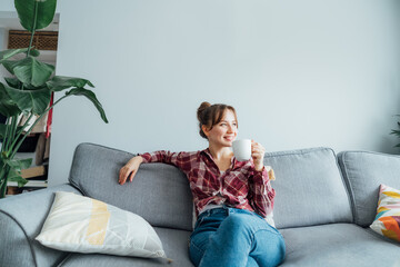 young smiling woman sitting on sofa and looking away while drinking coffee or tea. young brunette wo