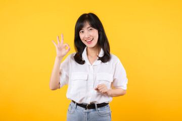 Wall Mural - Young businesswoman hand sign okay gesture. Asian woman happy smile wearing white shirt and jean denim plants showing okay sign hand gesture isolated on yellow background.