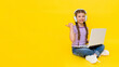 © Юлия Дьякова - A child with a laptop. A little girl sits on the floor and communicates online on a laptop, wearing headphones. Internet communication for children. Yellow isolated background. copy space.