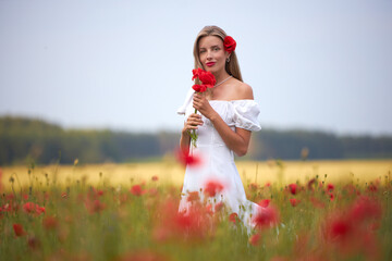 A young slim woman in a white dress walks in a flower field with poppies in summer.