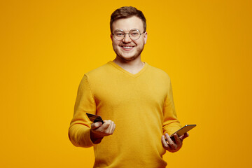 Young caucasian overjoyed bearded man wears orange sweater and glasses, holds smartphone and credit bank card isolated on orange background