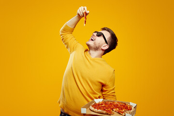 Excited man with sunglasses holding and biting tasty slice of pizza, posing with open mouth, holding cardboard flat box on yellow orange background