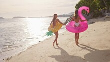 Two Girlfriends Walk Along The Beach At Sunset Holding Hands After Swimming In The Sea With Pineapple And Flamingo-shaped Inflatables. The Women Enjoy Their Vacation And Each Other's Time