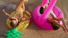 Two Girlfriends Wearing Sunglasses Lie On Inflatable Flamingo And Pineapple Shaped Circles And Talk. During A Weekend Vacation They Have A Good Time On The Beach.