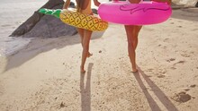 Two Girlfriends Walk Along The Beach At Sunset Holding Hands After Swimming In The Sea With Pineapple And Flamingo-shaped Inflatables. The Women Enjoy Their Vacation And Their Weekend