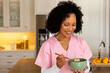 © wavebreak3 - Happy biracial female medical worker in scrubs eating bowl of breakfast cereal in kitchen at home