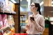 © _KUBE_ - Shopping. Side view portrait of pretty caucasian woman wearing glasses holds smartphone and choosing products in supermarket. Online communication and shopping