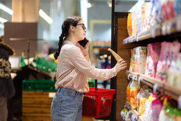 Wall Mural - Shopping. Pretty caucasian woman choosing food in supermarket and talking on smartphone. Side view