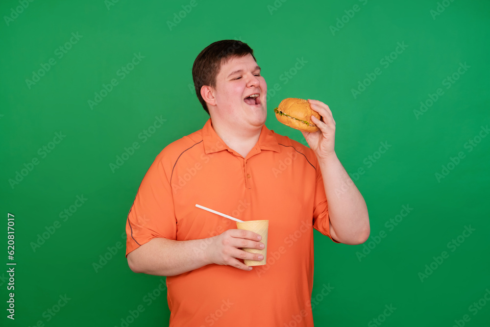 Portrait of a fat guy eating a big hamburger and drinking soda from a glass with a straw, isolated on a green background. Chroma key, green screen. The concept of fast food and obesity.
