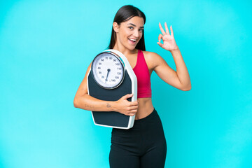 Wall Mural - Young caucasian woman isolated on blue background holding a weighing machine and doing OK sign