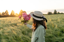 Woman Standing In A Wheat Field Holding A Bunch Of Pink Peonies, Belarus