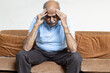 © Photographielove - Tired, depressed senior man sitting on couch in living room feeling hurt and lonely. Aged, white-haired man touching forehead suffering from severe headache or recalling bad memories