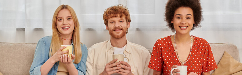 Wall Mural - polyamorous concept, relationship diversity, happy polygamy lovers  holding cups of coffee, looking at camera, interracial man and women in living room, bisexual and polygamy, banner