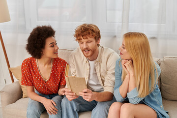 Wall Mural - open relationships, redhead man reading book to multicultural women in living room, non traditional lovers, cultural diversity, romance and love, modern family and polygamy concept