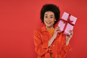 holiday, positive african american woman in casual attire holding wrapped present on coral background, vibrant colors, gift box, attractive and stylish, festive occasions, looking at camera