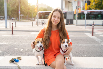 Smiling long haired Girl with two dog friends. Summer time vibe in big city. Smiling happy teenage girl in orange clothes hugging cute pets Jack Russells sitting on the bench. Public transport tram 