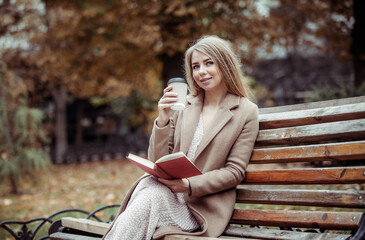 Cute beautiful woman with a cup of coffee and book sits on a bench in autumn park