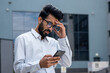 © Liubomir - Young hispanic businessman outside office building, man in shirt reading sad bad news online using app on phone, depressed man disappointed unhappy with achievement results.