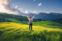 Happy Girl On The Hill With Yellow Flowers And Green Grass In Beautiful Alpine Mountain Valley At Sunset In Summer. Landscape With Young Woman With Raised Up Arms In Alps, Trees, Sky With Clouds
