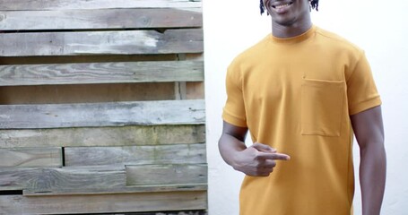 Wall Mural - Happy african american man wearing yellow t shirt pointing to pocket, slow motion, copy space