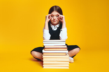 Wall Mural - Shocked schoolgirl open eyes with fingers and looking at bunch of books, not ready for studying, sitting isolated over yellow background