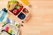 © photoguns - School lunch box with sandwich, fruits, vegetables, bottle of water, and kids backpack with school supplies on wooden table. Flat lay, top view.