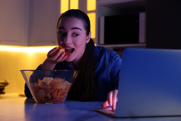 Wall Mural - Young woman eating chips while using laptop in kitchen at night. Bad habit