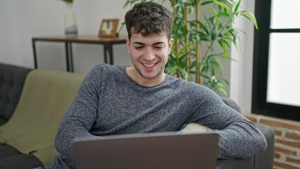 Wall Mural - Young hispanic man using laptop sitting on sofa at home