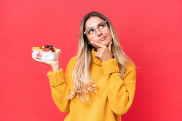 Wall Mural - Young pretty Uruguayan woman holding a bowl of fruit isolated on red background having doubts while looking up