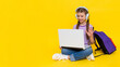 © Юлия Дьякова - The child is engaged in an online lesson with a laptop. A young girl is sitting cross-legged on the floor with a backpack and headphones. Yellow isolated background. Copy space.