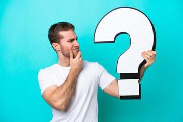 Young caucasian man isolated on blue background holding a question mark icon and having doubts