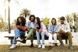 © carlesiturbe - Multiethnic group of friends texting on their smartphones sitting on a bench in the street. Diverse young people smiling and using their phones outside in a park bench.
