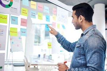 Creative man, sticky note and pointing on glass board for schedule tasks, planning or brainstorming at office. Male person or employee with plan, agenda or marketing strategy for startup at workplace