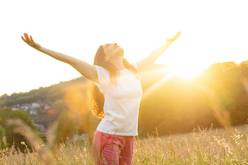 A woman stands in a field in nature with her hands raised in the rays of the setting sun in summer.