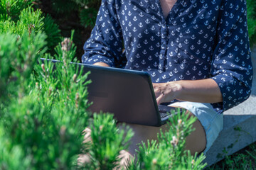 Woman freelance works on laptop in city park. Young adult student education in fresh air. Female sits outdoor and types on computer. Workplace on nature in sunny day. Distance online 5g internet.