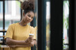 © Songsak C - A smiling black woman drinking coffee while standing near a window at a cafe, a Happy young African American female holding a cup with a hot drink and looking away.