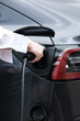 © Treerat - Young woman traveling by electric car having stop at charging station standing plugging charger close-up.
