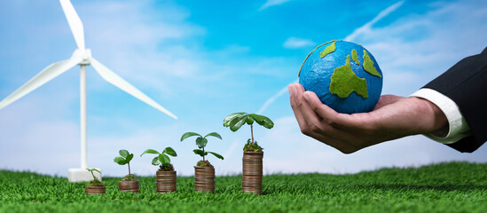 Eco business invest on environment conservation subsidize, coin stack with grow seedling, hand holding paper Earth and wind turbine. Sustainable financial growth with clean and renewable energy. Alter