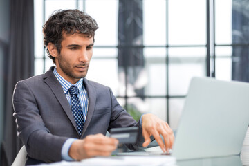 Smiling man sitting in office and pays by credit card with his laptop.