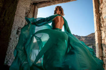 Rear view of a happy blonde woman in a long mint dress posing against the backdrop of the sea in an old building with columns. Girl in nature against the blue sky.