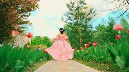 Wall Mural - Fantasy girl princess runs in spring garden flowers tulips tree green grass alley. Woman queen, old style long pink dress with train skirt fabric fly in wind slow motion. fairy lady back rear view 4k