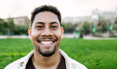 Outside smiling portrait of young latino hispanic gen z man looking at camera standing at city park. Front headshot of joyful student guy laughing outdoors.