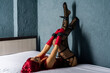 Slave red-haired woman in a corset and red leather gloves seductively posing lying back on the bed and legs up.