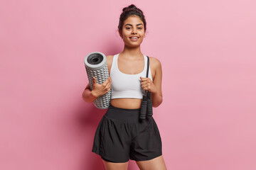 Studio shot of cheerful sporty young woman smiles gladfully holds massage roller and jumping rope dressed in cropped white t shirt and black shorts isolated over pink background. Sport concept