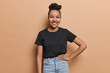 Horizontal shot of pretty young Latin woman with hair bun keeps hand on waist smiles pleasantly stands self confident wears casual black t shirt and fashionable jeans isolated over brown background.